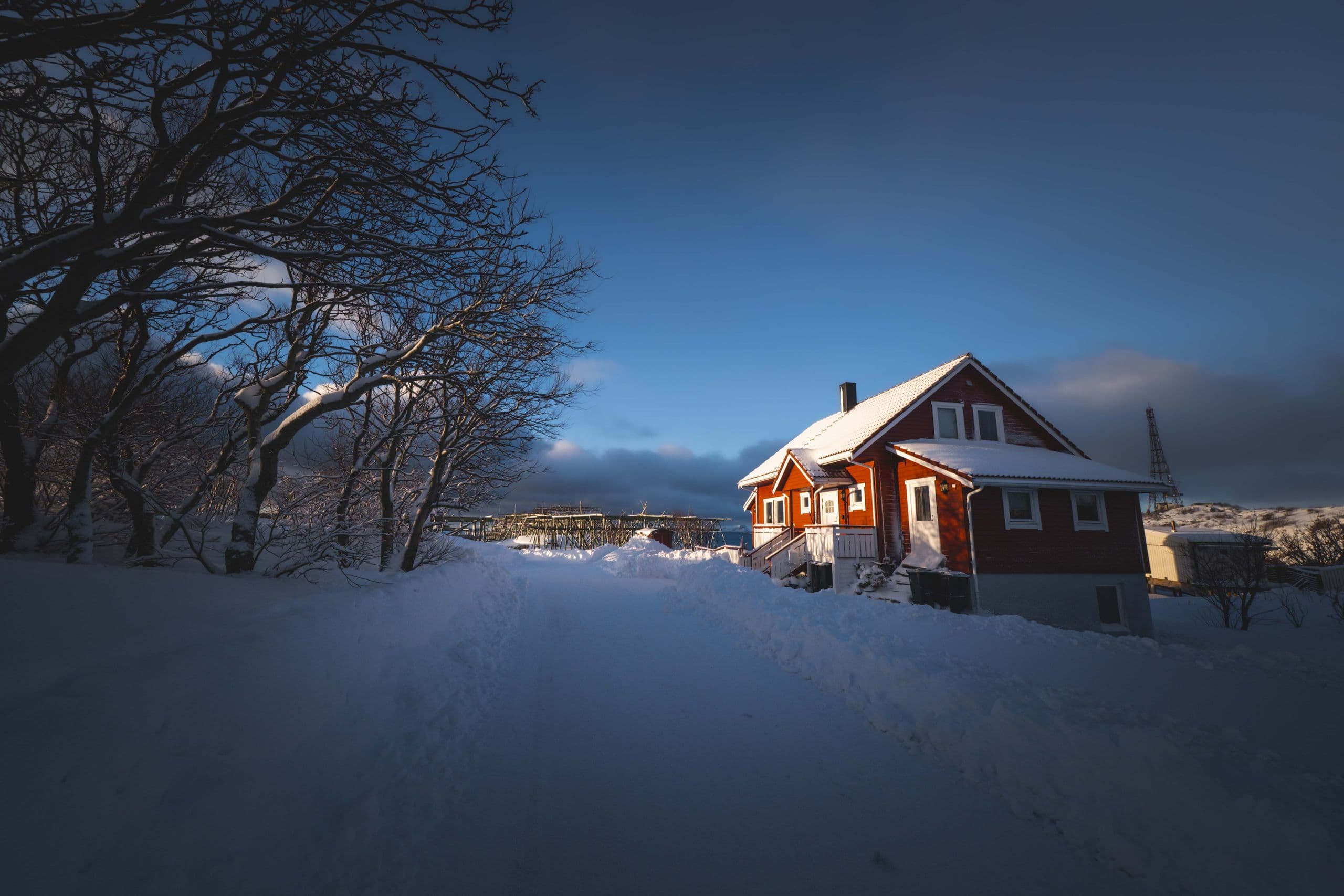 Red house in deep snow at dusk with open snow and bare trees to its left